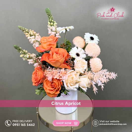 Citrus Apricot vase arrangement featuring orange and peach roses, white gerberas, orange sprayed pompons, snapdragons, dried ruscus, hydra, and sinamomo styled in a modern vase.